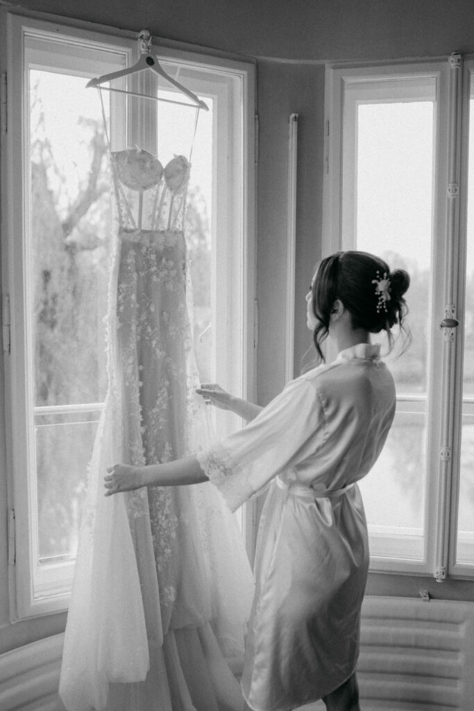 Braut betrachtet ihr Hochzeitskleid vor Fenster