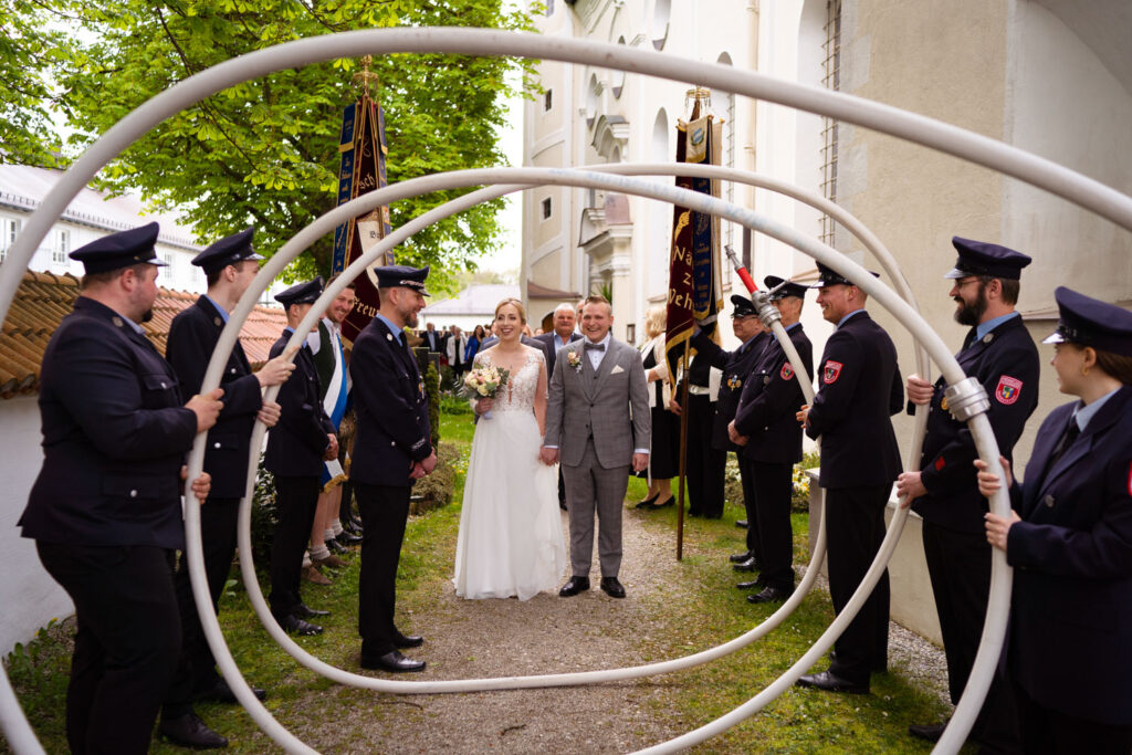 Hochzeitspaar unter Feuerwehrschläuchen vor Kirche feiernd