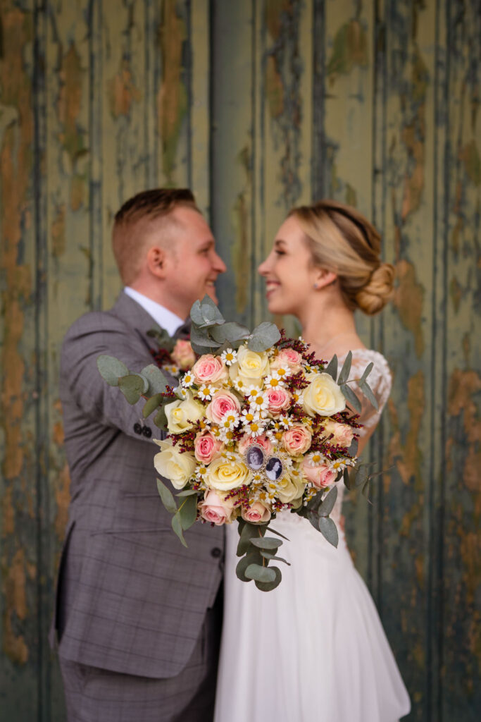 Lächelndes Brautpaar mit Blumenstrauß vor rustikaler Wand